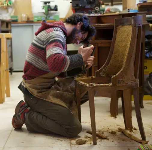 A craftsman kneeling to restore an antique wooden chair